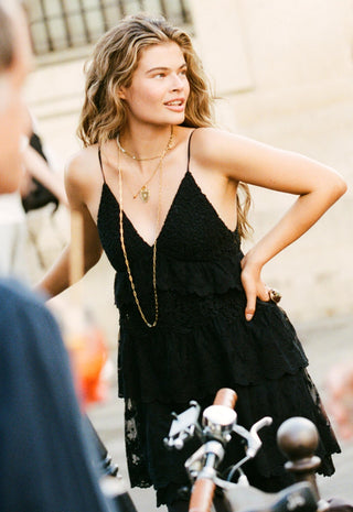 a-woman-wearing-a-black-lace-tiered-spaghetti-strap-dress-and-gold-necklaces-standing-outdoors-next-to-a-bicycle