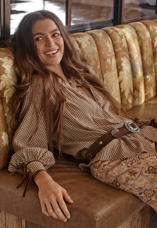 A woman with long brown hair reclines on a brown leather bench wearing a brown and beige gingham long-sleeve top with a matching brown belt and patterned pants, set against a rustic background with a patterned gold and brown velvet sofa.